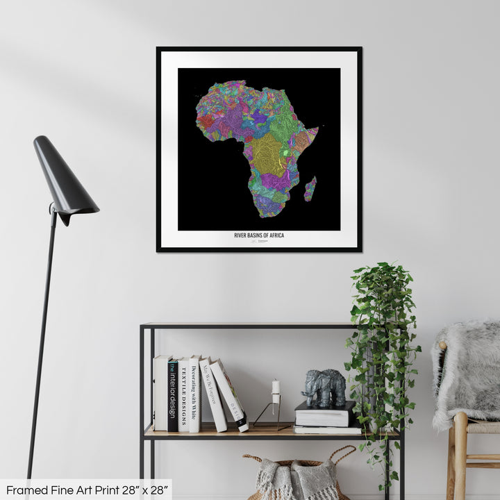 A colourful Grasshopper Geography map on a white wall, home decor in a living room. The map is in a black frame with a thick white mat. The title of the map is printed on the mat. The map hangs above a black shelf with plants and books on. A black lamp stands in the corner.