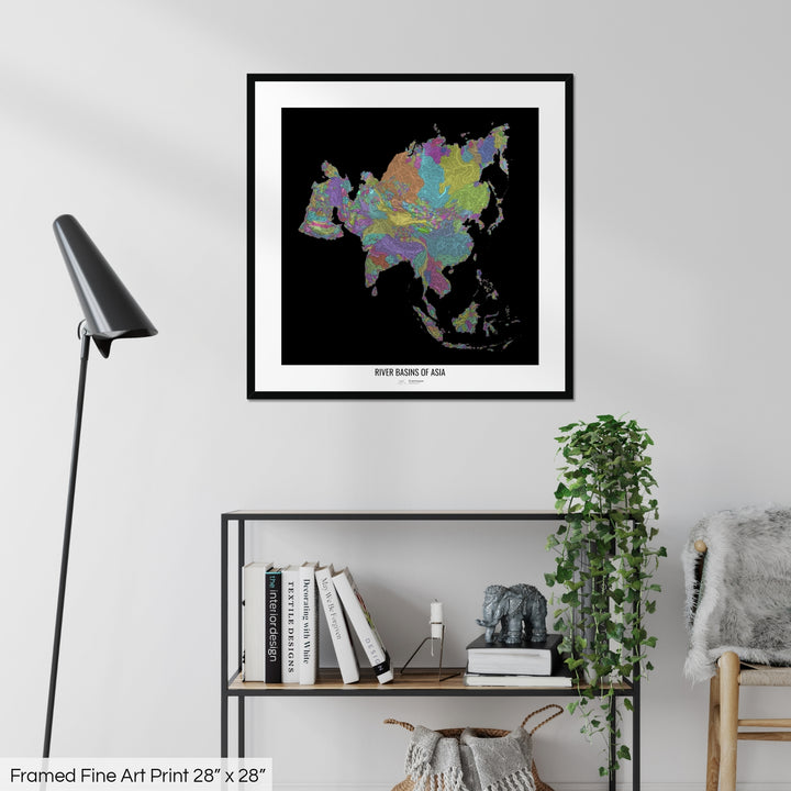 A colourful Grasshopper Geography map on a white wall, home decor in a living room. The map is in a black frame with a thick white mat. The title of the map is printed on the mat. The map hangs above a black shelf with plants and books on. A black lamp stands in the corner.