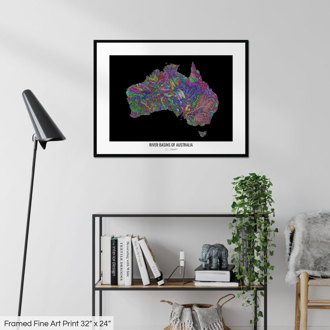 A colourful Grasshopper Geography map on a white wall, home decor in a living room. The map is in a black frame with a thick white mat. The title of the map is printed on the mat. The map hangs above a black shelf with plants and books on. A black lamp stands in the corner.
