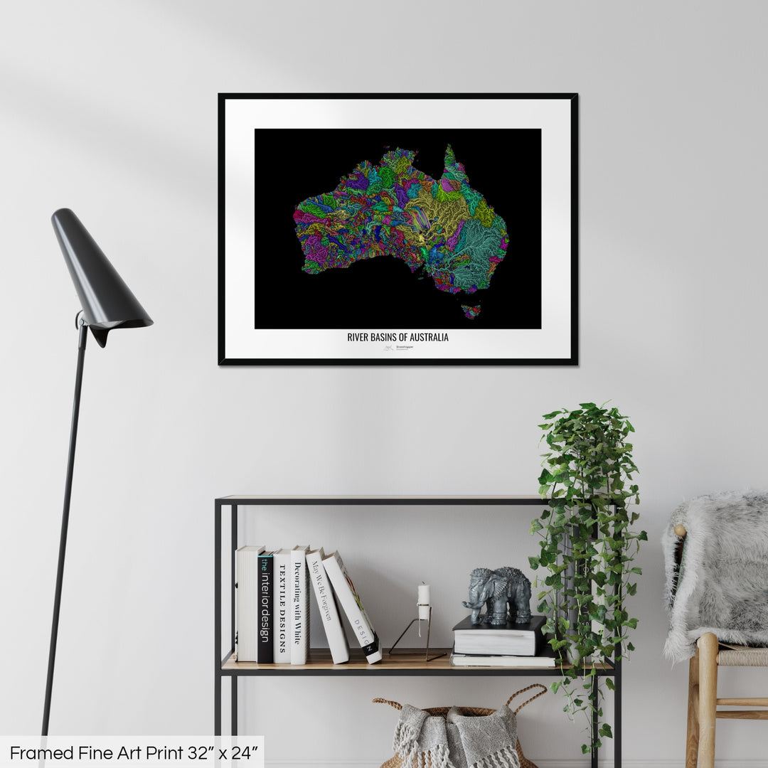 A colourful Grasshopper Geography map on a white wall, home decor in a living room. The map is in a black frame with a thick white mat. The title of the map is printed on the mat. The map hangs above a black shelf with plants and books on. A black lamp stands in the corner.