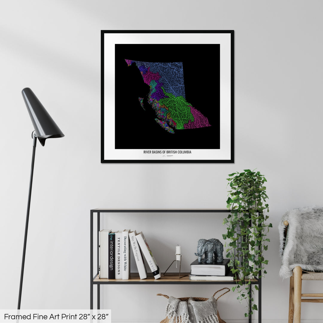A colourful Grasshopper Geography map on a white wall, home decor in a living room. The map is in a black frame with a thick white mat. The title of the map is printed on the mat. The map hangs above a black shelf with plants and books on. A black lamp stands in the corner.