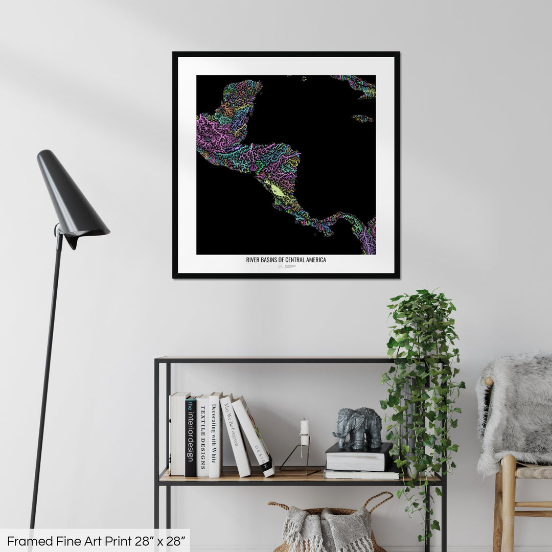 A colourful Grasshopper Geography map on a white wall, home decor in a living room. The map is in a black frame with a thick white mat. The title of the map is printed on the mat. The map hangs above a black shelf with plants and books on. A black lamp stands in the corner.