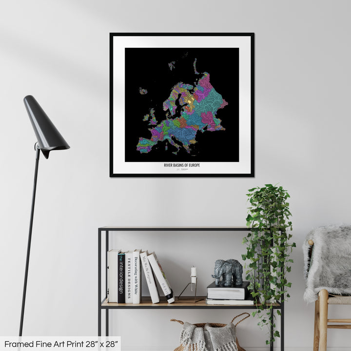 A colourful Grasshopper Geography map on a white wall, home decor in a living room. The map is in a black frame with a thick white mat. The title of the map is printed on the mat. The map hangs above a black shelf with plants and books on. A black lamp stands in the corner.