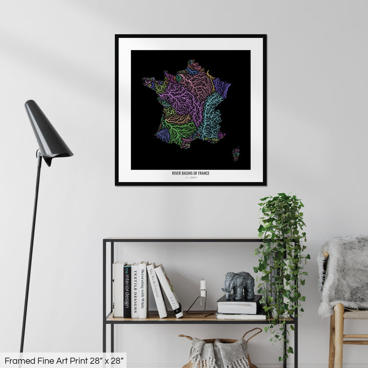 A colourful Grasshopper Geography map on a white wall, home decor in a living room. The map is in a black frame with a thick white mat. The title of the map is printed on the mat. The map hangs above a black shelf with plants and books on. A black lamp stands in the corner.
