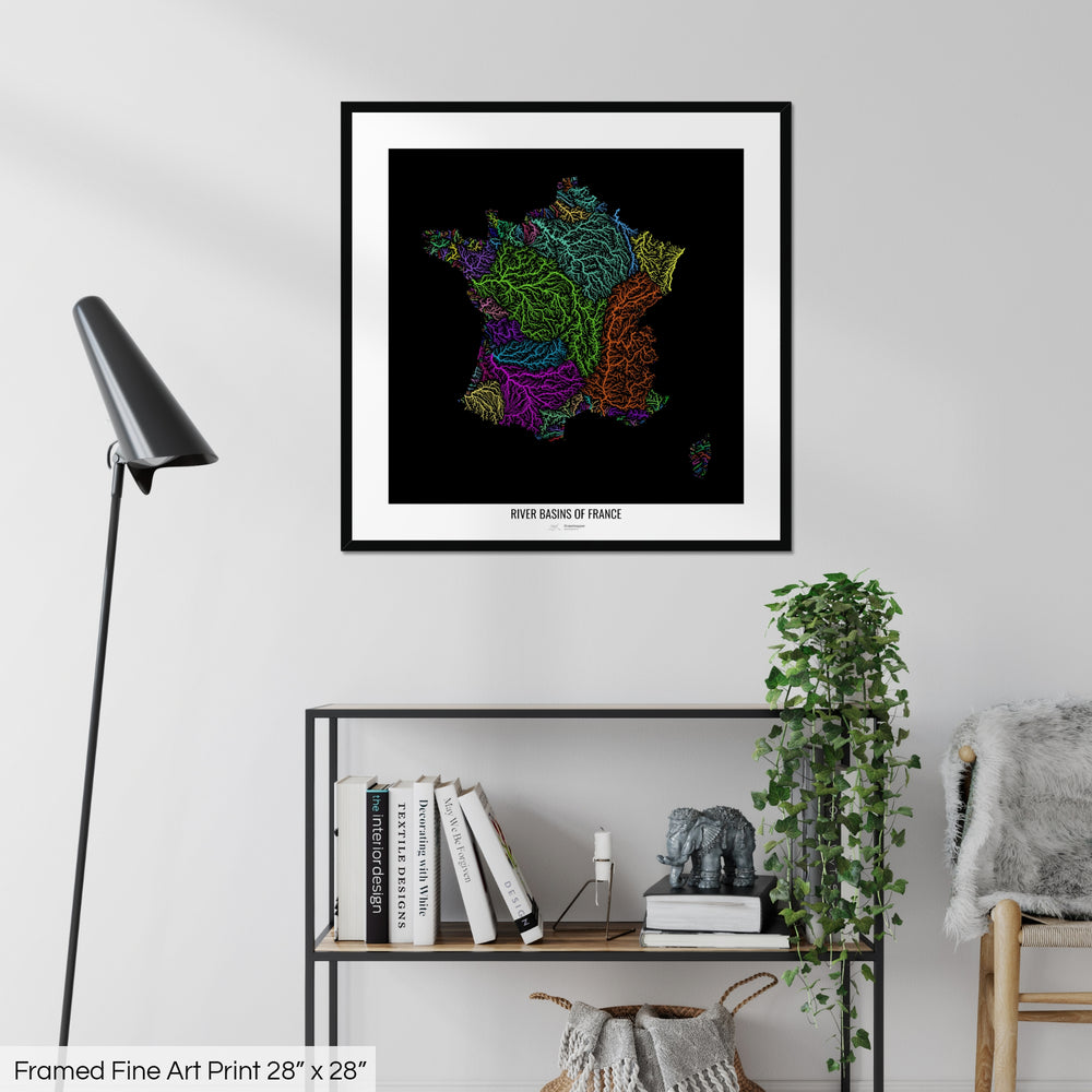 A colourful Grasshopper Geography map on a white wall, home decor in a living room. The map is in a black frame with a thick white mat. The title of the map is printed on the mat. The map hangs above a black shelf with plants and books on. A black lamp stands in the corner.