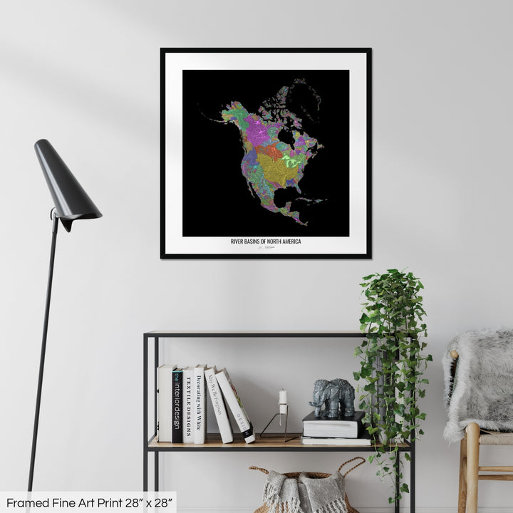 A colourful Grasshopper Geography map on a white wall, home decor in a living room. The map is in a black frame with a thick white mat. The title of the map is printed on the mat. The map hangs above a black shelf with plants and books on. A black lamp stands in the corner.