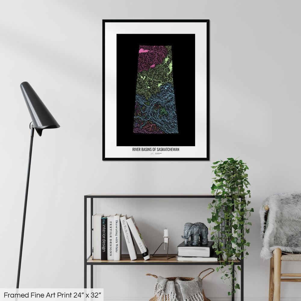 A colourful Grasshopper Geography map on a white wall, home decor in a living room. The map is in a black frame with a thick white mat. The title of the map is printed on the mat. The map hangs above a black shelf with plants and books on. A black lamp stands in the corner.