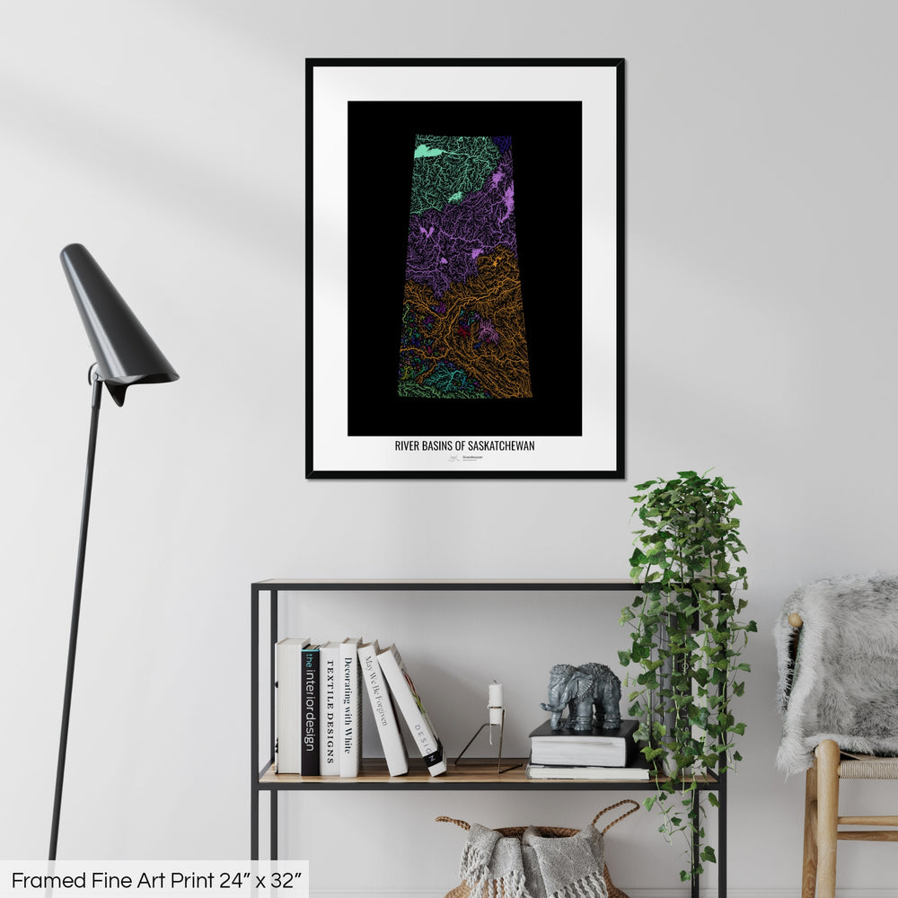 A colourful Grasshopper Geography map on a white wall, home decor in a living room. The map is in a black frame with a thick white mat. The title of the map is printed on the mat. The map hangs above a black shelf with plants and books on. A black lamp stands in the corner.