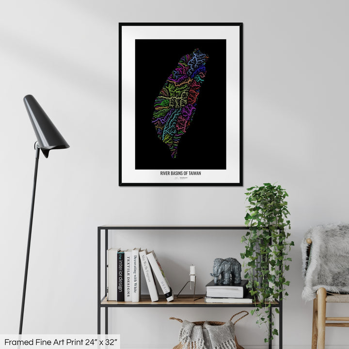 A colourful Grasshopper Geography map on a white wall, home decor in a living room. The map is in a black frame with a thick white mat. The title of the map is printed on the mat. The map hangs above a black shelf with plants and books on. A black lamp stands in the corner.