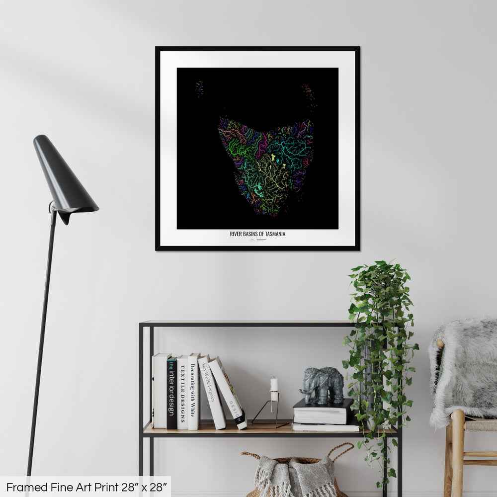 A colourful Grasshopper Geography map on a white wall, home decor in a living room. The map is in a black frame with a thick white mat. The title of the map is printed on the mat. The map hangs above a black shelf with plants and books on. A black lamp stands in the corner.