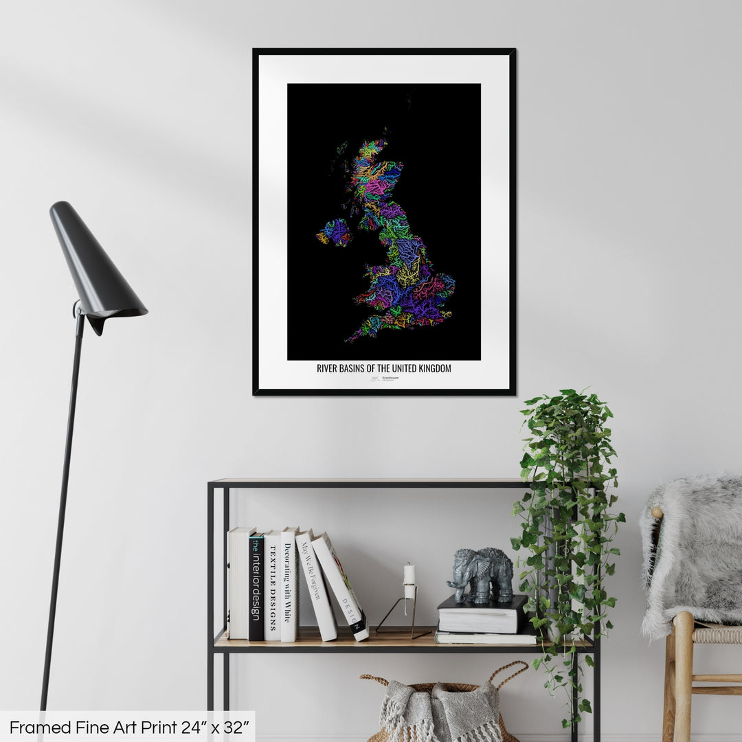 A colourful Grasshopper Geography map on a white wall, home decor in a living room. The map is in a black frame with a thick white mat. The title of the map is printed on the mat. The map hangs above a black shelf with plants and books on. A black lamp stands in the corner.