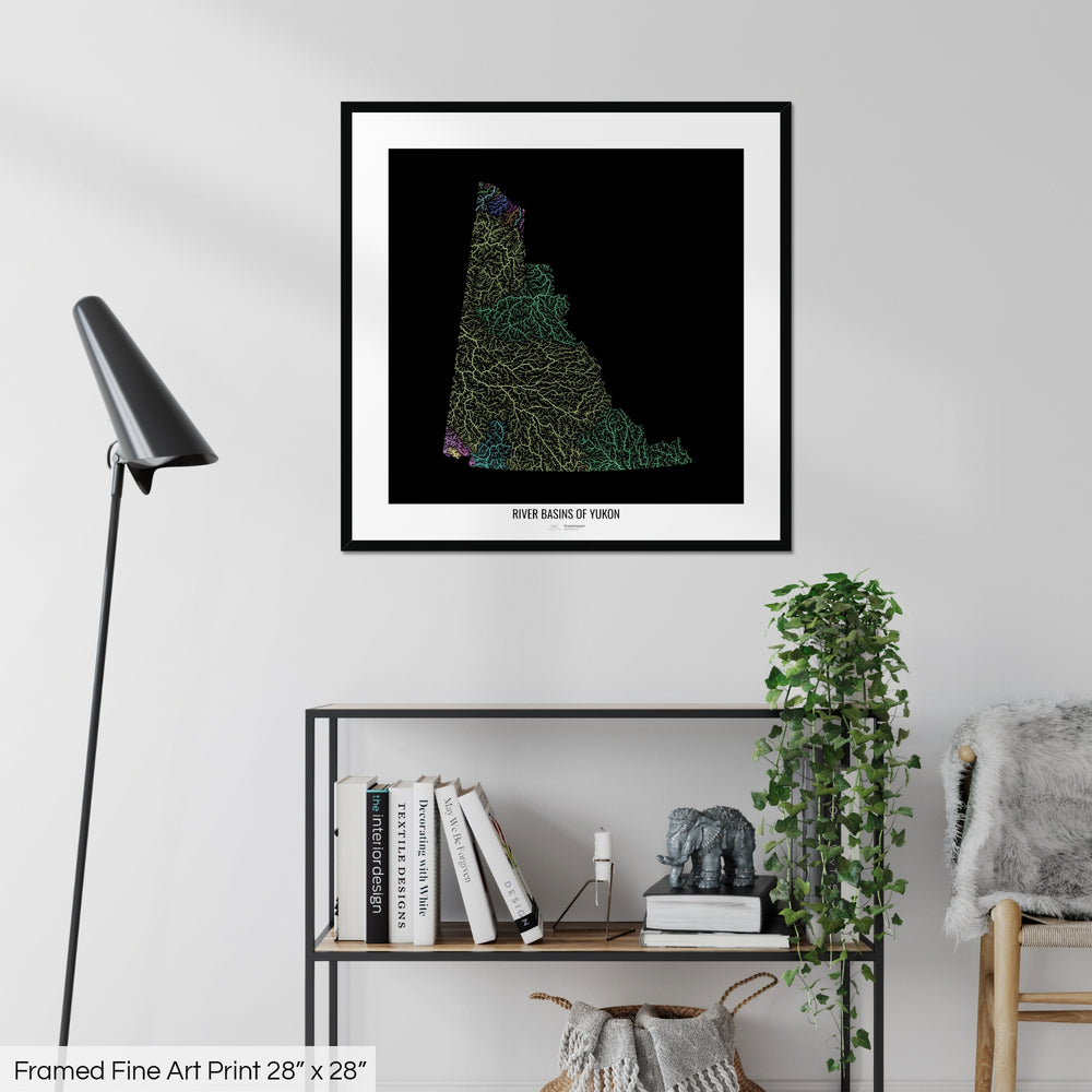 A colourful Grasshopper Geography map on a white wall, home decor in a living room. The map is in a black frame with a thick white mat. The title of the map is printed on the mat. The map hangs above a black shelf with plants and books on. A black lamp stands in the corner.