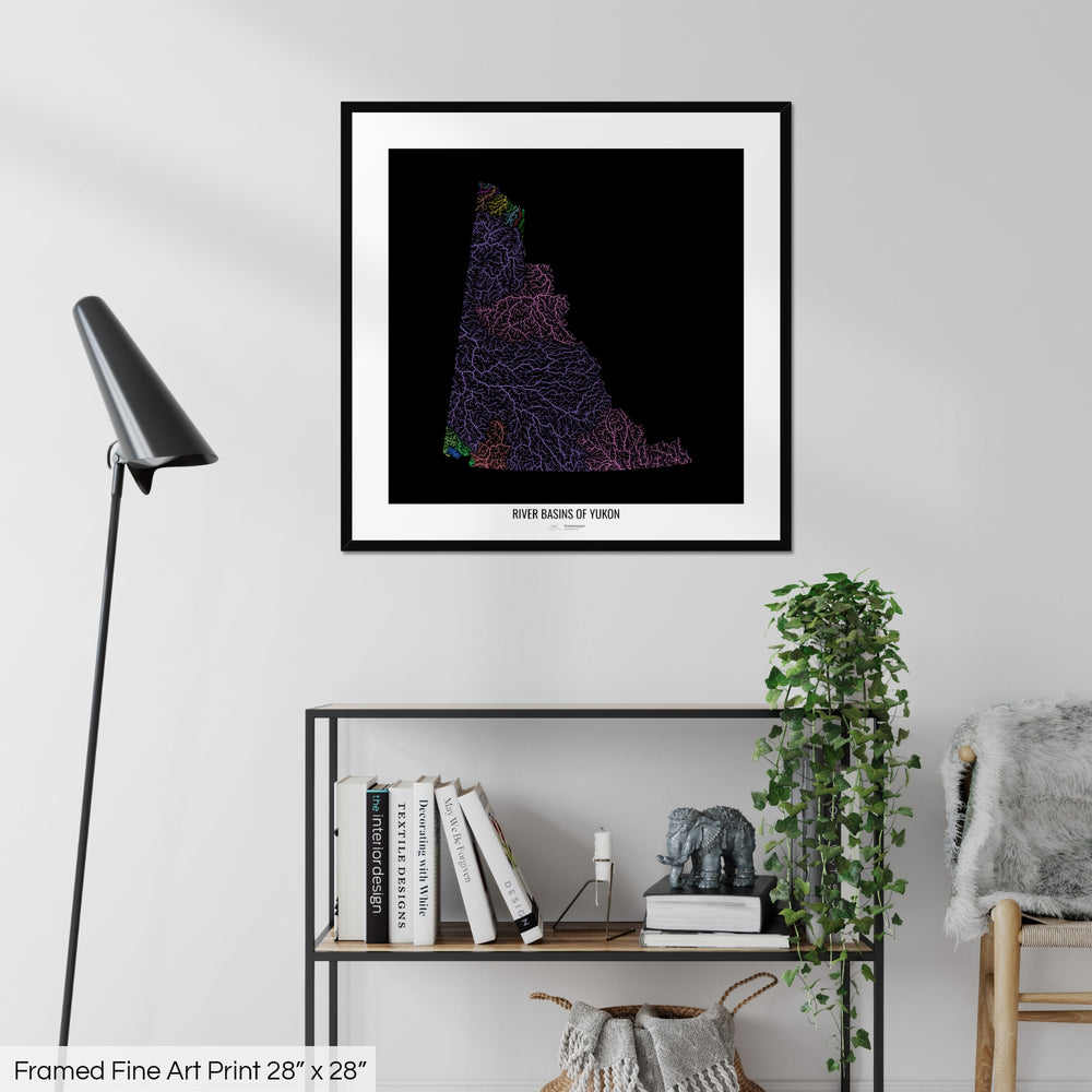 A colourful Grasshopper Geography map on a white wall, home decor in a living room. The map is in a black frame with a thick white mat. The title of the map is printed on the mat. The map hangs above a black shelf with plants and books on. A black lamp stands in the corner.