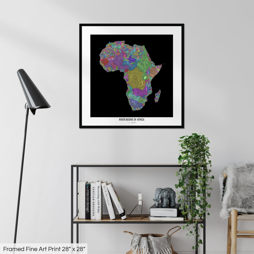 A colourful Grasshopper Geography map on a white wall, home decor in a living room. The map is in a black frame with a thick white mat. The title of the map is printed on the mat. The map hangs above a black shelf with plants and books on. A black lamp stands in the corner.
