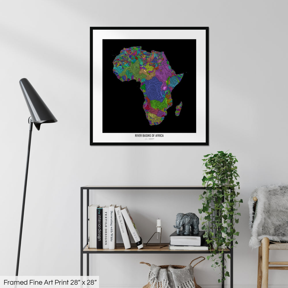 A colourful Grasshopper Geography map on a white wall, home decor in a living room. The map is in a black frame with a thick white mat. The title of the map is printed on the mat. The map hangs above a black shelf with plants and books on. A black lamp stands in the corner.