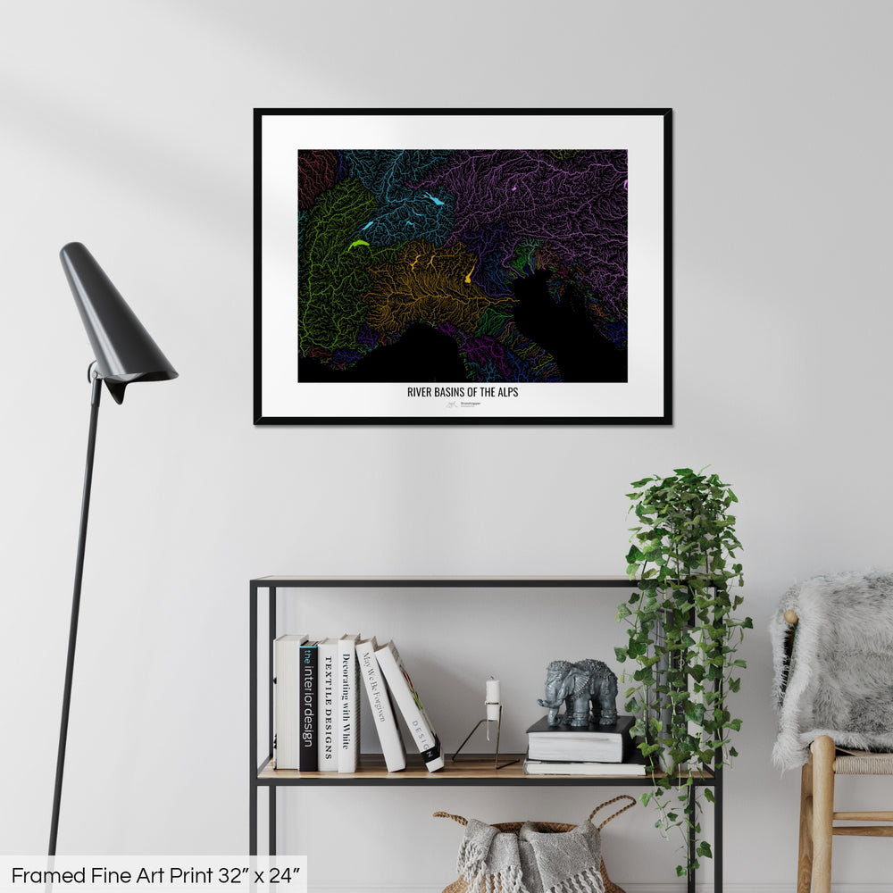 A colourful Grasshopper Geography map on a white wall, home decor in a living room. The map is in a black frame with a thick white mat. The title of the map is printed on the mat. The map hangs above a black shelf with plants and books on. A black lamp stands in the corner.