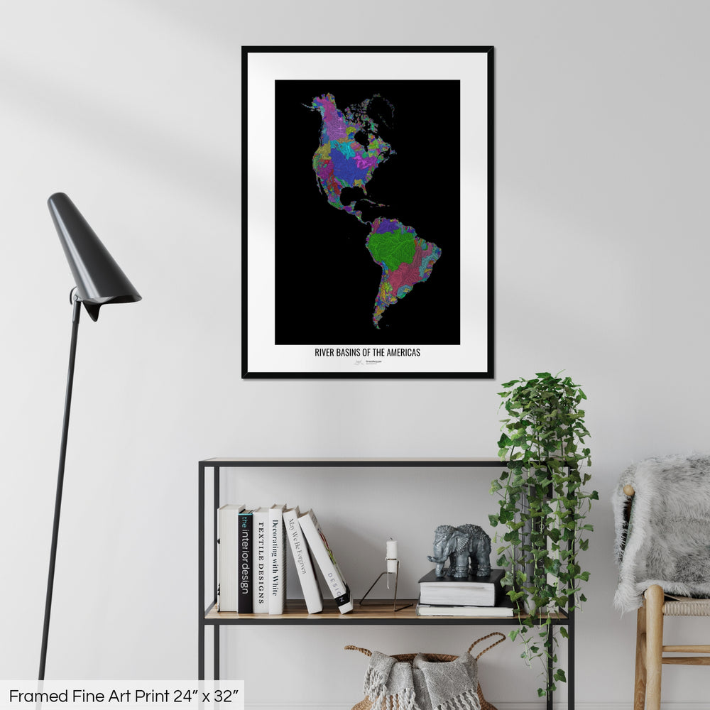 A colourful Grasshopper Geography map on a white wall, home decor in a living room. The map is in a black frame with a thick white mat. The title of the map is printed on the mat. The map hangs above a black shelf with plants and books on. A black lamp stands in the corner.