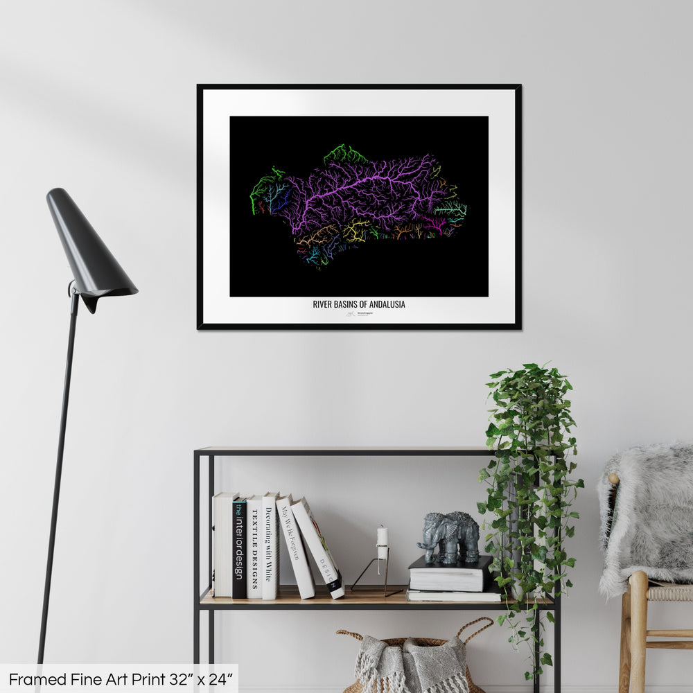 A colourful Grasshopper Geography map on a white wall, home decor in a living room. The map is in a black frame with a thick white mat. The title of the map is printed on the mat. The map hangs above a black shelf with plants and books on. A black lamp stands in the corner.