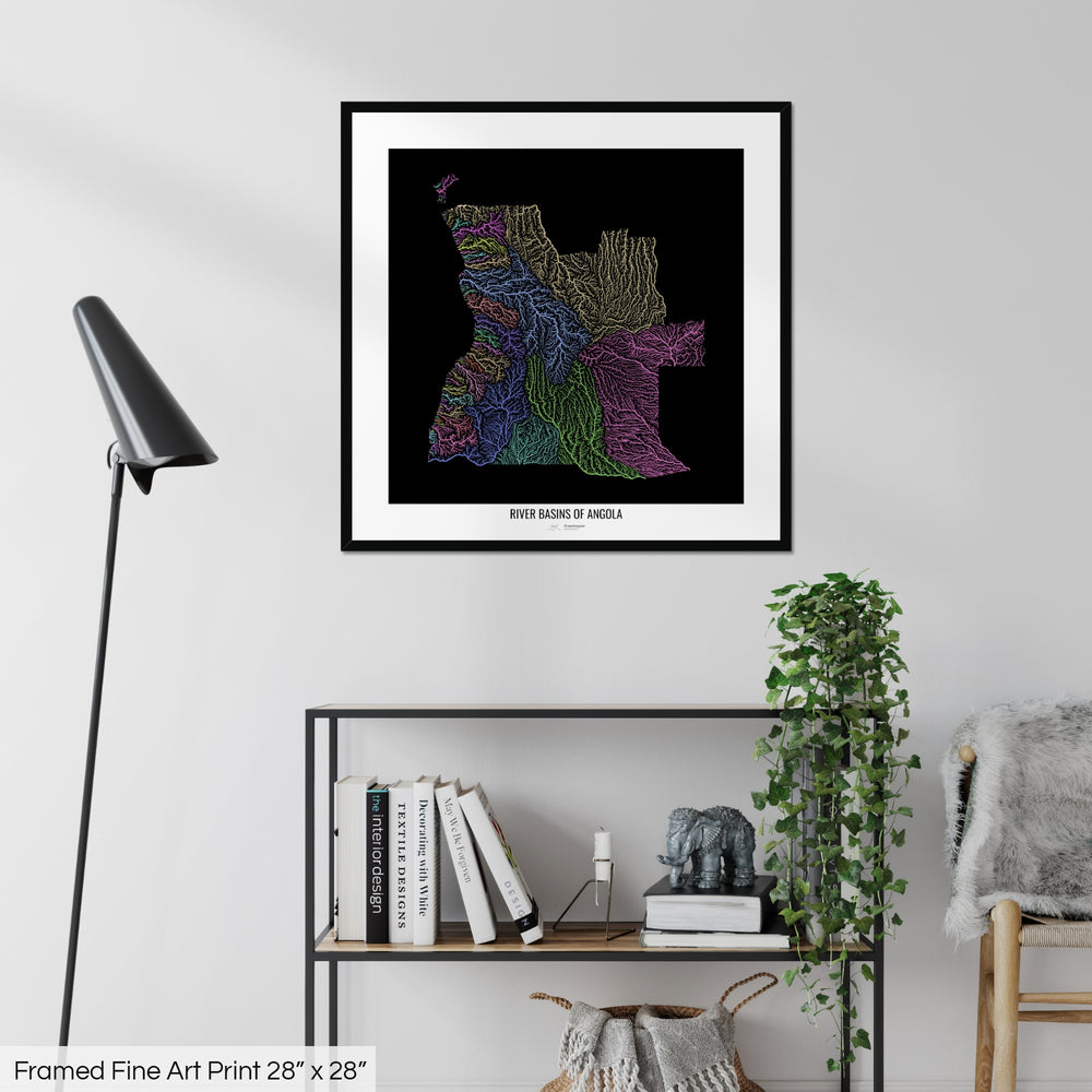 A colourful Grasshopper Geography map on a white wall, home decor in a living room. The map is in a black frame with a thick white mat. The title of the map is printed on the mat. The map hangs above a black shelf with plants and books on. A black lamp stands in the corner.