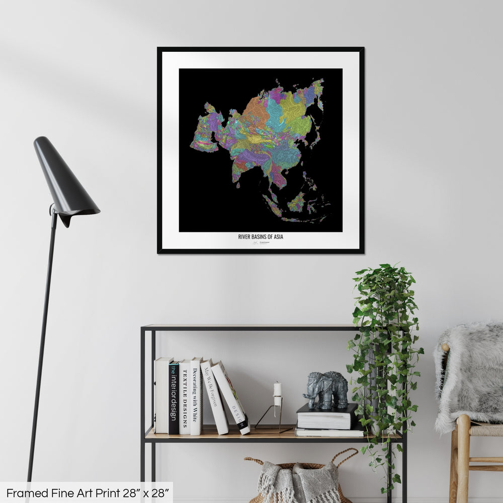 A colourful Grasshopper Geography map on a white wall, home decor in a living room. The map is in a black frame with a thick white mat. The title of the map is printed on the mat. The map hangs above a black shelf with plants and books on. A black lamp stands in the corner.