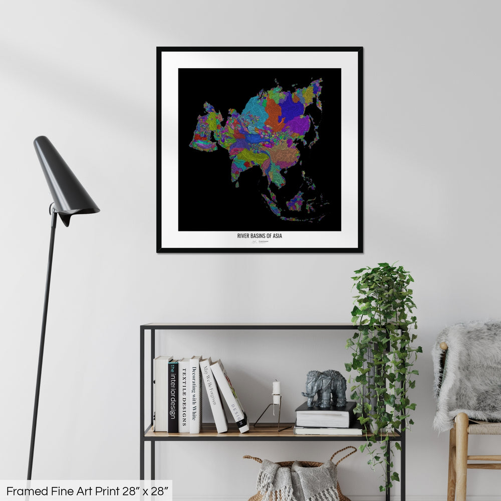 A colourful Grasshopper Geography map on a white wall, home decor in a living room. The map is in a black frame with a thick white mat. The title of the map is printed on the mat. The map hangs above a black shelf with plants and books on. A black lamp stands in the corner.