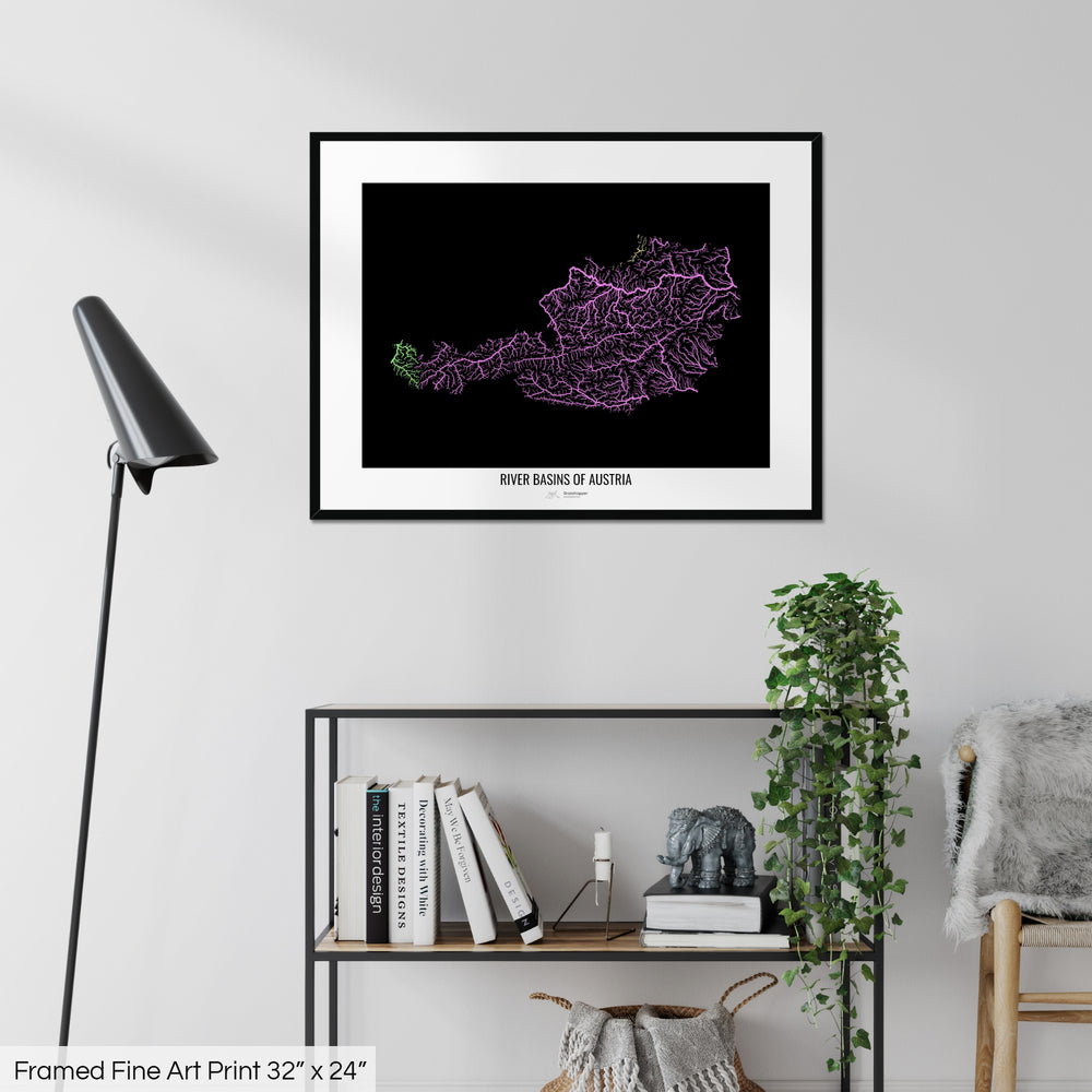 A colourful Grasshopper Geography map on a white wall, home decor in a living room. The map is in a black frame with a thick white mat. The title of the map is printed on the mat. The map hangs above a black shelf with plants and books on. A black lamp stands in the corner.