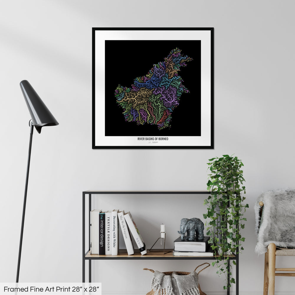 A colourful Grasshopper Geography map on a white wall, home decor in a living room. The map is in a black frame with a thick white mat. The title of the map is printed on the mat. The map hangs above a black shelf with plants and books on. A black lamp stands in the corner.