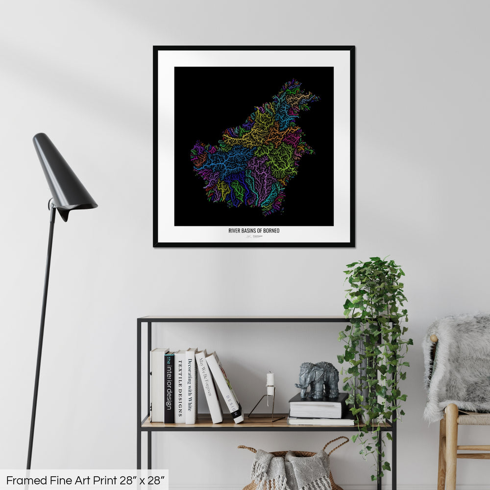 A colourful Grasshopper Geography map on a white wall, home decor in a living room. The map is in a black frame with a thick white mat. The title of the map is printed on the mat. The map hangs above a black shelf with plants and books on. A black lamp stands in the corner.