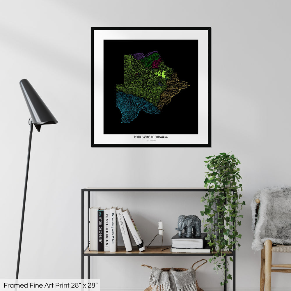 A colourful Grasshopper Geography map on a white wall, home decor in a living room. The map is in a black frame with a thick white mat. The title of the map is printed on the mat. The map hangs above a black shelf with plants and books on. A black lamp stands in the corner.