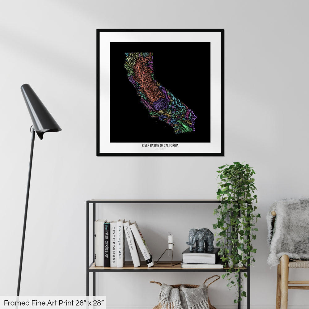 A colourful Grasshopper Geography map on a white wall, home decor in a living room. The map is in a black frame with a thick white mat. The title of the map is printed on the mat. The map hangs above a black shelf with plants and books on. A black lamp stands in the corner.
