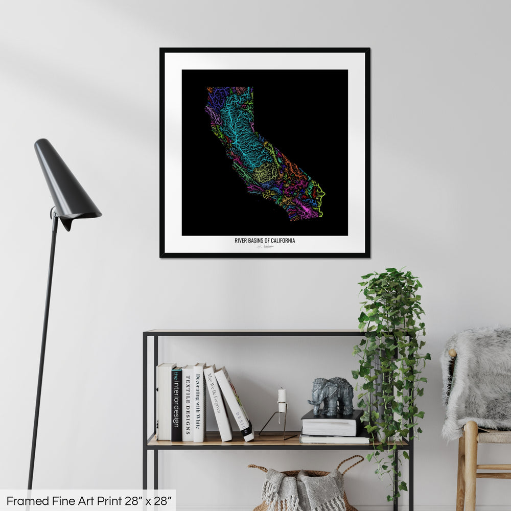 A colourful Grasshopper Geography map on a white wall, home decor in a living room. The map is in a black frame with a thick white mat. The title of the map is printed on the mat. The map hangs above a black shelf with plants and books on. A black lamp stands in the corner.