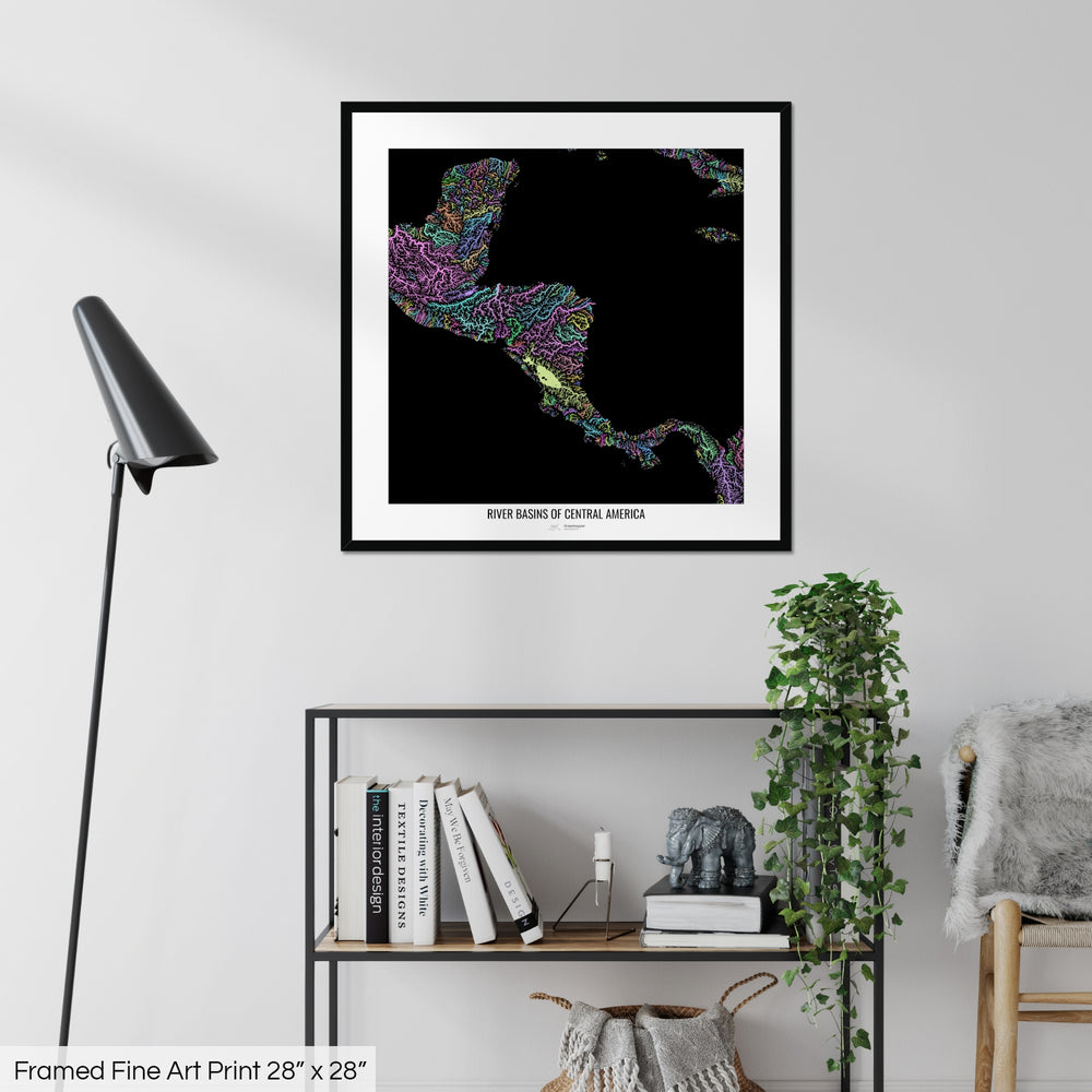 A colourful Grasshopper Geography map on a white wall, home decor in a living room. The map is in a black frame with a thick white mat. The title of the map is printed on the mat. The map hangs above a black shelf with plants and books on. A black lamp stands in the corner.
