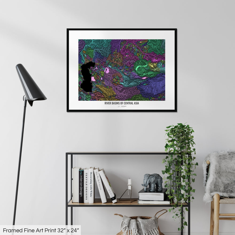 A colourful Grasshopper Geography map on a white wall, home decor in a living room. The map is in a black frame with a thick white mat. The title of the map is printed on the mat. The map hangs above a black shelf with plants and books on. A black lamp stands in the corner.