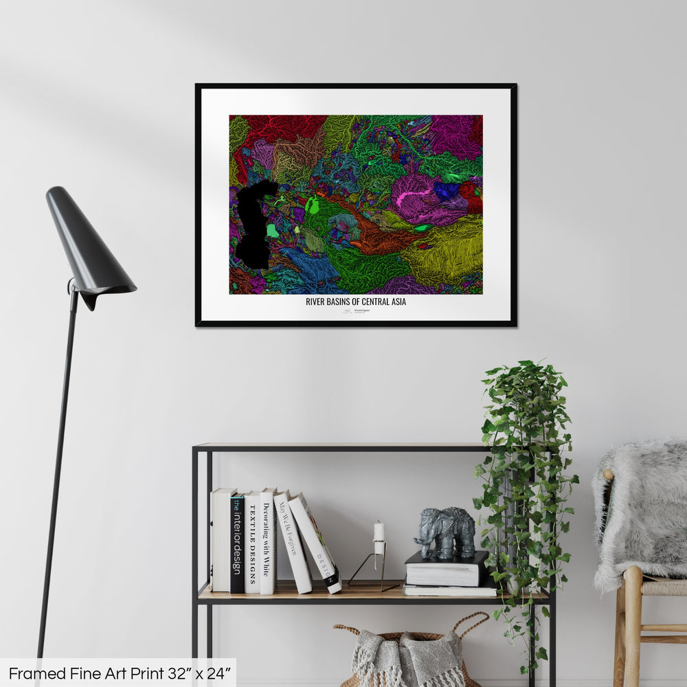 A colourful Grasshopper Geography map on a white wall, home decor in a living room. The map is in a black frame with a thick white mat. The title of the map is printed on the mat. The map hangs above a black shelf with plants and books on. A black lamp stands in the corner.