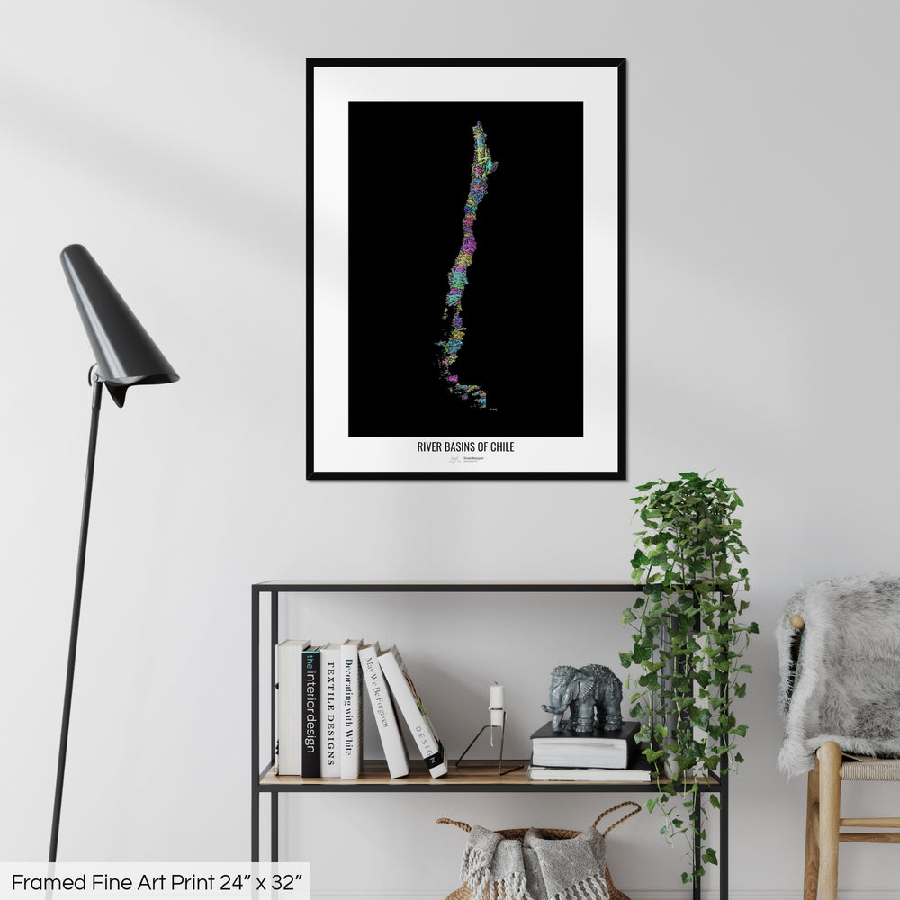 A colourful Grasshopper Geography map on a white wall, home decor in a living room. The map is in a black frame with a thick white mat. The title of the map is printed on the mat. The map hangs above a black shelf with plants and books on. A black lamp stands in the corner.