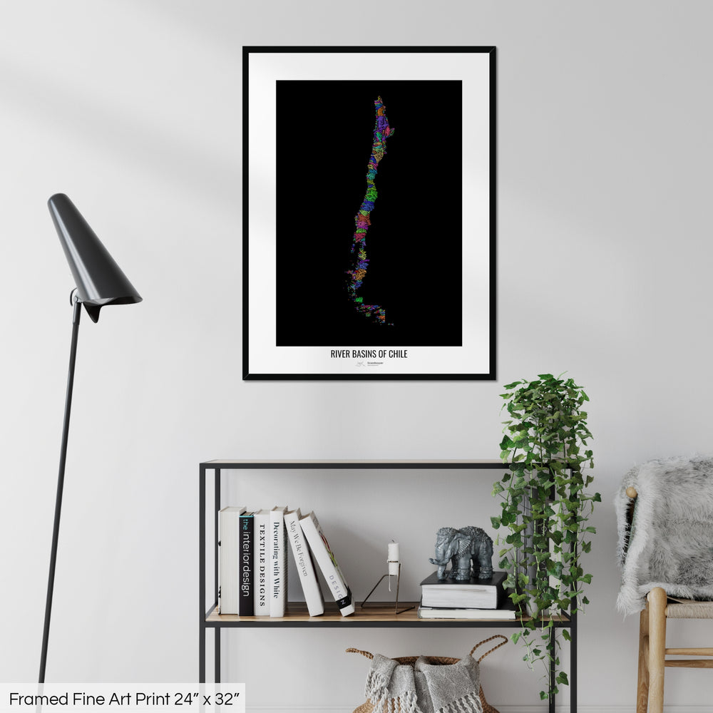 A colourful Grasshopper Geography map on a white wall, home decor in a living room. The map is in a black frame with a thick white mat. The title of the map is printed on the mat. The map hangs above a black shelf with plants and books on. A black lamp stands in the corner.