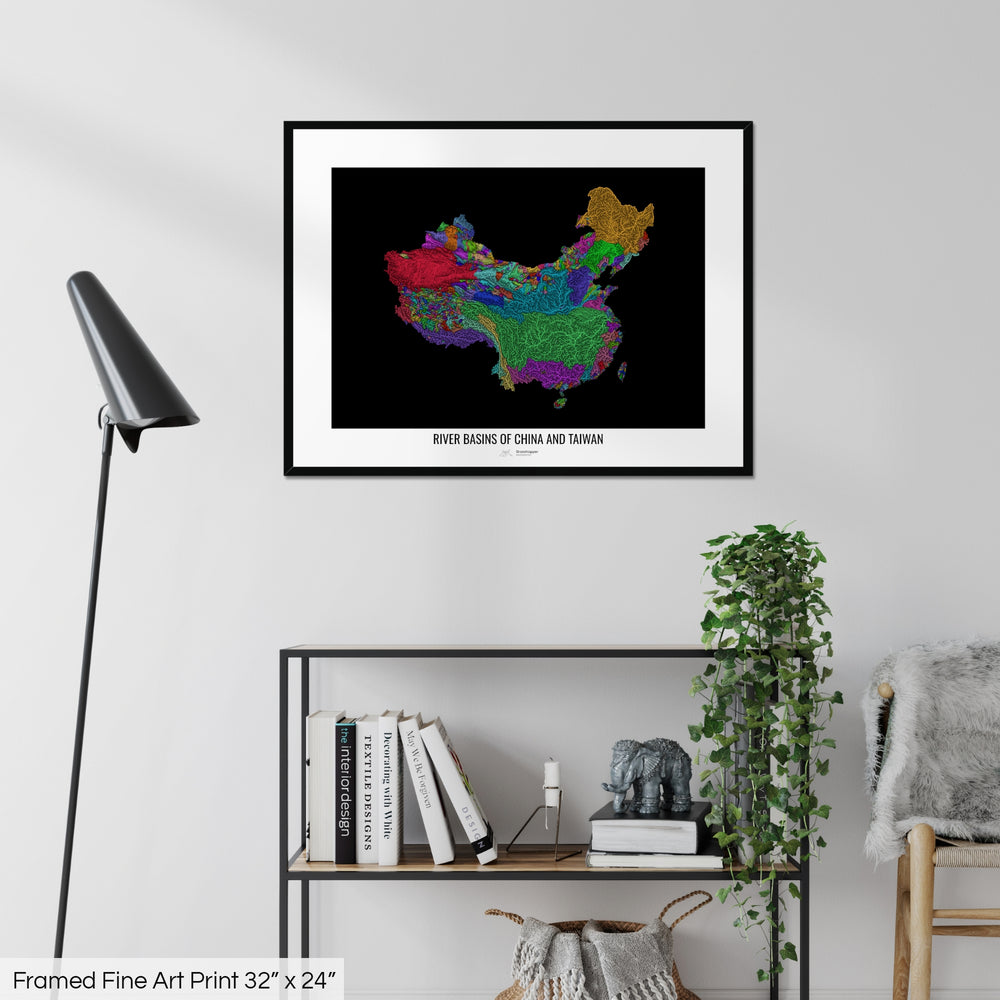 A colourful Grasshopper Geography map on a white wall, home decor in a living room. The map is in a black frame with a thick white mat. The title of the map is printed on the mat. The map hangs above a black shelf with plants and books on. A black lamp stands in the corner.