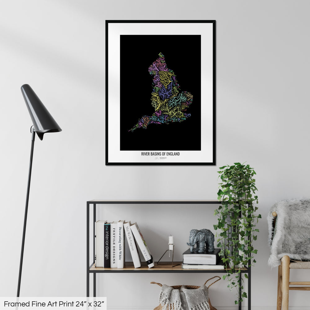 A colourful Grasshopper Geography map on a white wall, home decor in a living room. The map is in a black frame with a thick white mat. The title of the map is printed on the mat. The map hangs above a black shelf with plants and books on. A black lamp stands in the corner.