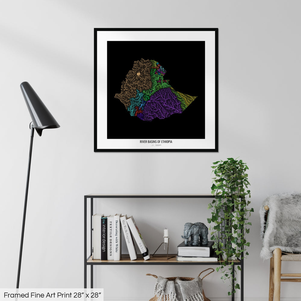 A colourful Grasshopper Geography map on a white wall, home decor in a living room. The map is in a black frame with a thick white mat. The title of the map is printed on the mat. The map hangs above a black shelf with plants and books on. A black lamp stands in the corner.