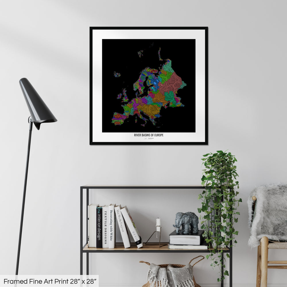 A colourful Grasshopper Geography map on a white wall, home decor in a living room. The map is in a black frame with a thick white mat. The title of the map is printed on the mat. The map hangs above a black shelf with plants and books on. A black lamp stands in the corner.