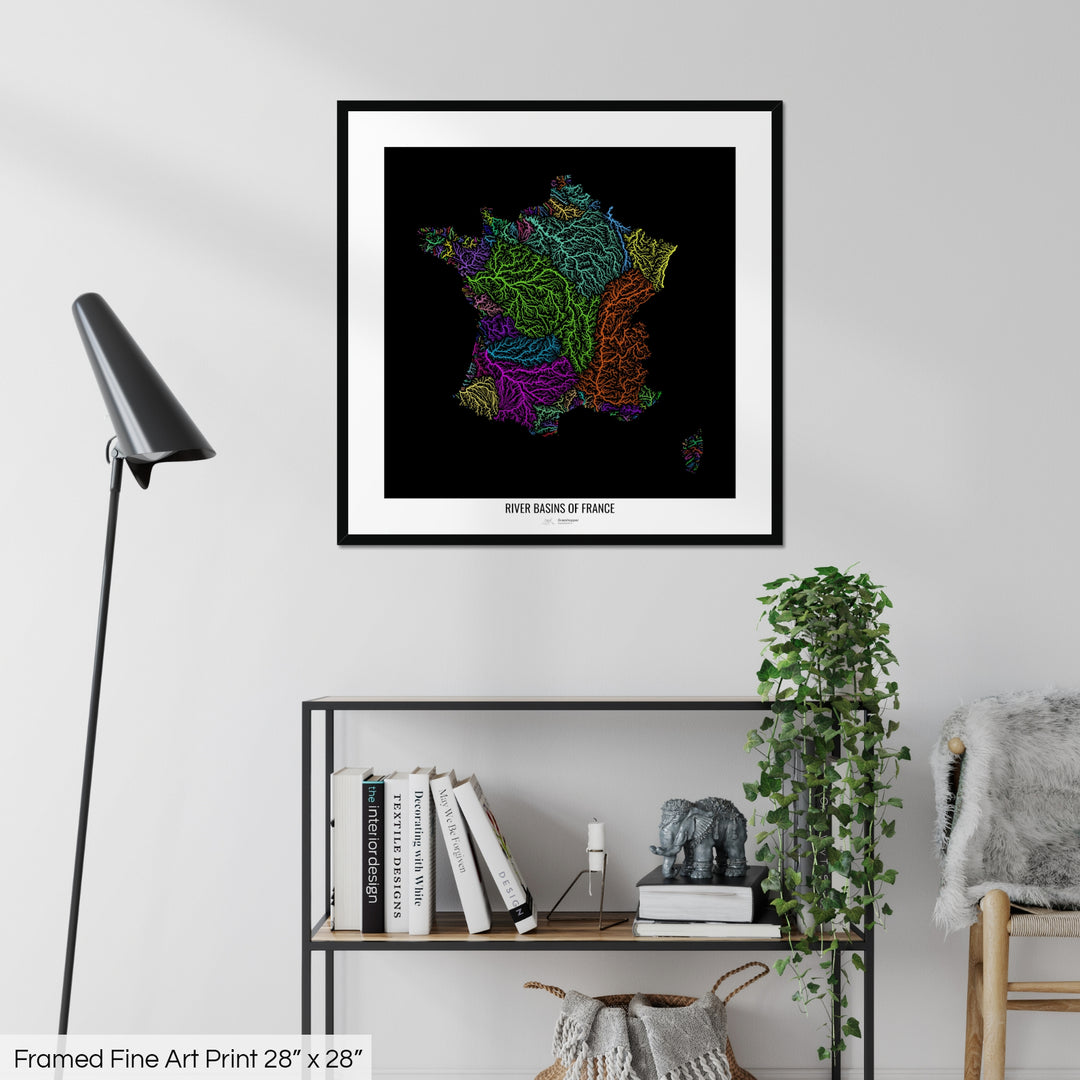 A colourful Grasshopper Geography map on a white wall, home decor in a living room. The map is in a black frame with a thick white mat. The title of the map is printed on the mat. The map hangs above a black shelf with plants and books on. A black lamp stands in the corner.