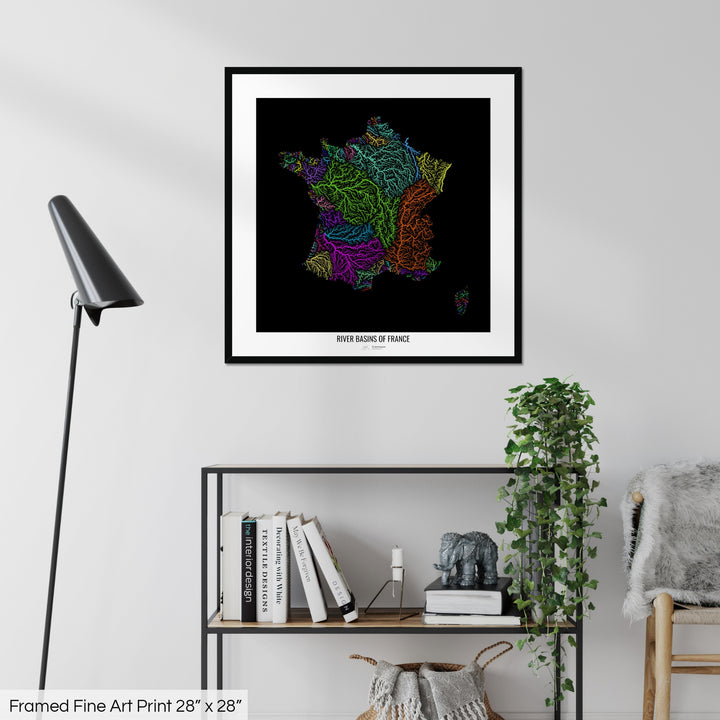 A colourful Grasshopper Geography map on a white wall, home decor in a living room. The map is in a black frame with a thick white mat. The title of the map is printed on the mat. The map hangs above a black shelf with plants and books on. A black lamp stands in the corner.