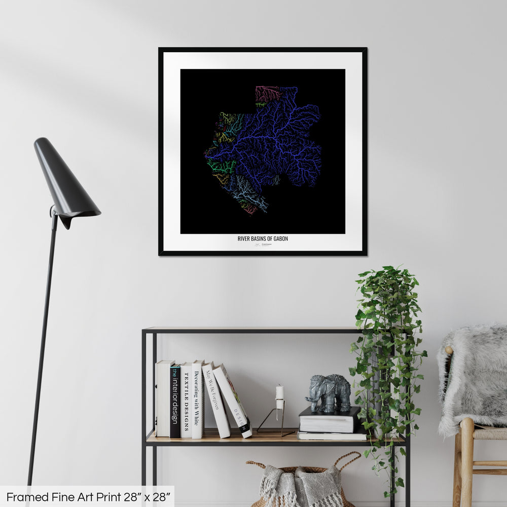 A colourful Grasshopper Geography map on a white wall, home decor in a living room. The map is in a black frame with a thick white mat. The title of the map is printed on the mat. The map hangs above a black shelf with plants and books on. A black lamp stands in the corner.