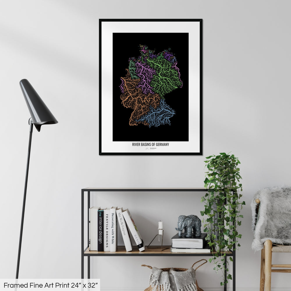 A colourful Grasshopper Geography map on a white wall, home decor in a living room. The map is in a black frame with a thick white mat. The title of the map is printed on the mat. The map hangs above a black shelf with plants and books on. A black lamp stands in the corner.