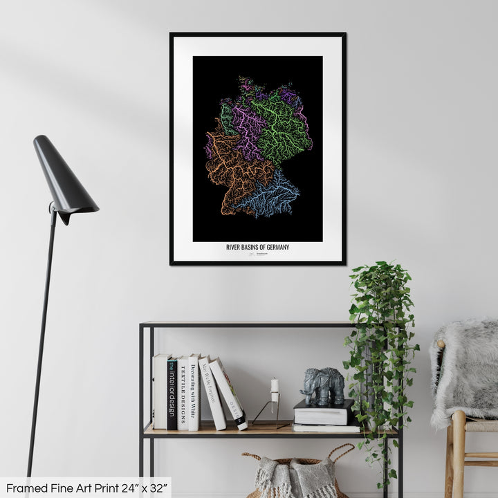 A colourful Grasshopper Geography map on a white wall, home decor in a living room. The map is in a black frame with a thick white mat. The title of the map is printed on the mat. The map hangs above a black shelf with plants and books on. A black lamp stands in the corner.
