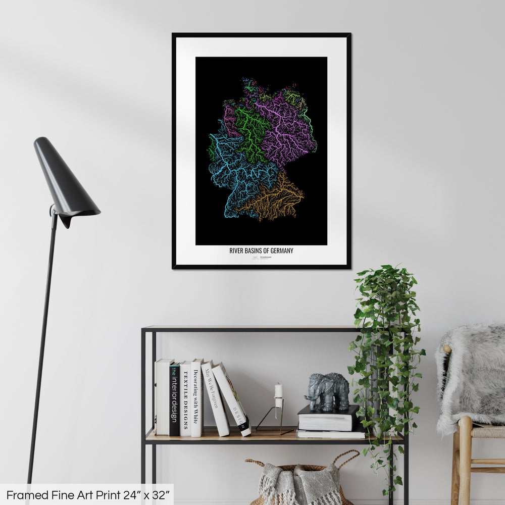 A colourful Grasshopper Geography map on a white wall, home decor in a living room. The map is in a black frame with a thick white mat. The title of the map is printed on the mat. The map hangs above a black shelf with plants and books on. A black lamp stands in the corner.
