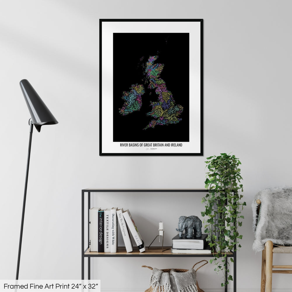 A colourful Grasshopper Geography map on a white wall, home decor in a living room. The map is in a black frame with a thick white mat. The title of the map is printed on the mat. The map hangs above a black shelf with plants and books on. A black lamp stands in the corner.