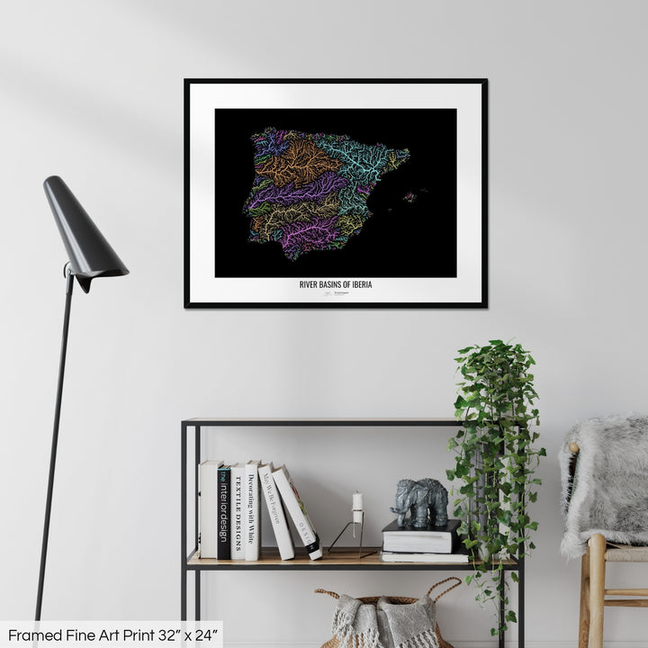 A colourful Grasshopper Geography map on a white wall, home decor in a living room. The map is in a black frame with a thick white mat. The title of the map is printed on the mat. The map hangs above a black shelf with plants and books on. A black lamp stands in the corner.
