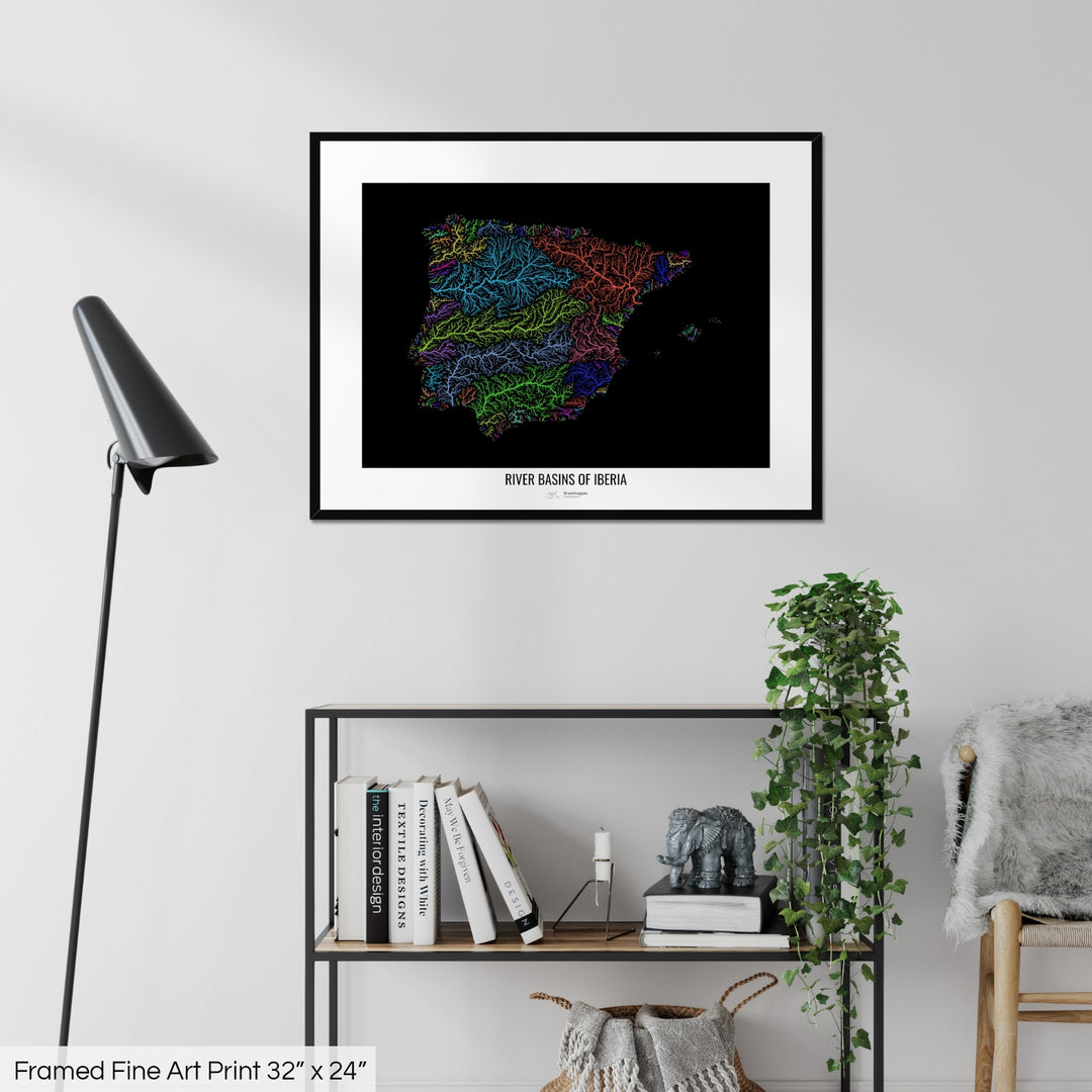 A colourful Grasshopper Geography map on a white wall, home decor in a living room. The map is in a black frame with a thick white mat. The title of the map is printed on the mat. The map hangs above a black shelf with plants and books on. A black lamp stands in the corner.