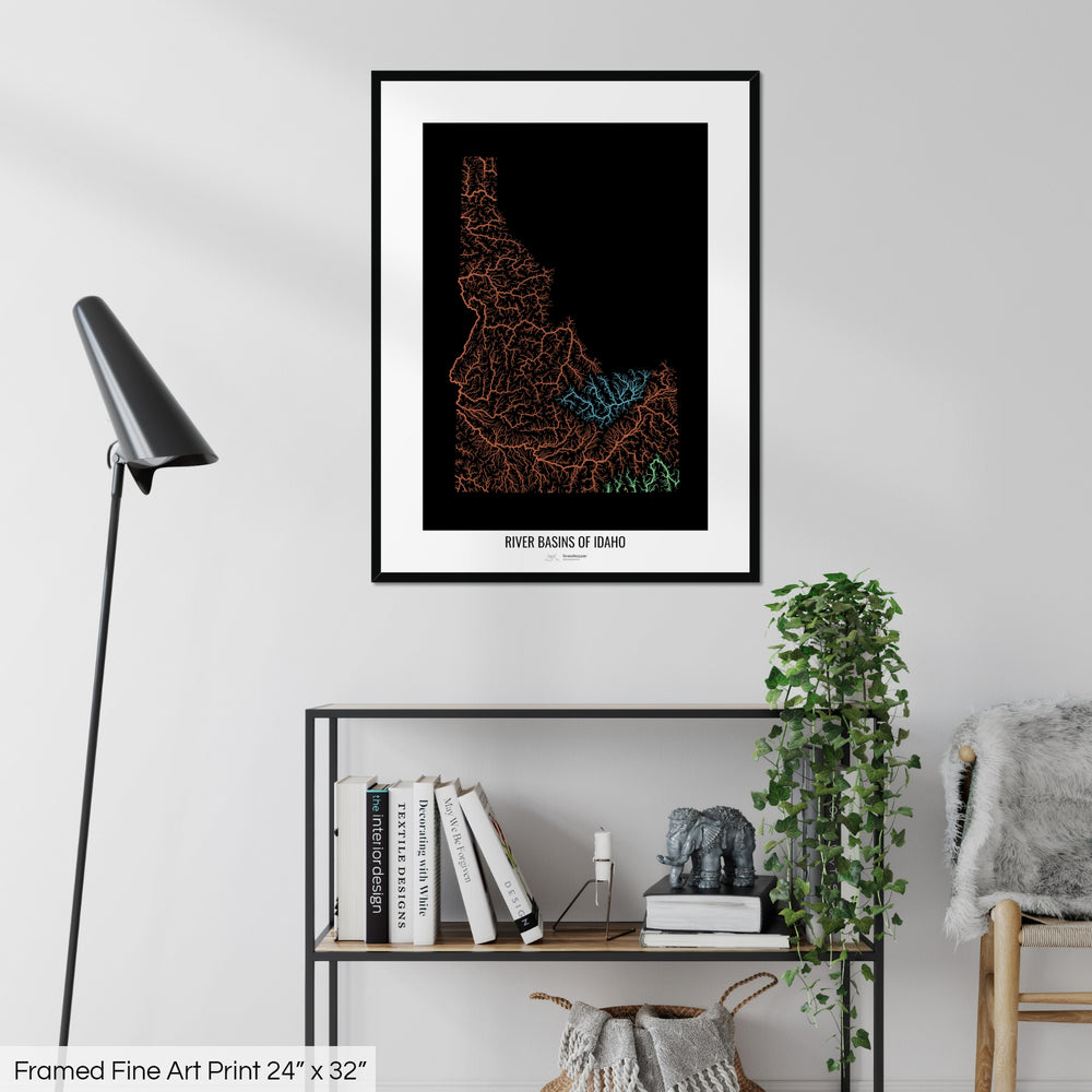 A colourful Grasshopper Geography map on a white wall, home decor in a living room. The map is in a black frame with a thick white mat. The title of the map is printed on the mat. The map hangs above a black shelf with plants and books on. A black lamp stands in the corner.
